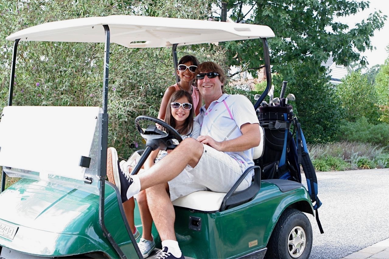 A dad and two kids in sunglasses smiling from a green golf cart during the neighborhood tournament