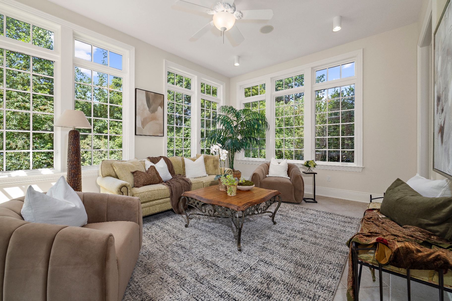 Current sunroom with cream sofa, brown accent chairs, wrought-iron coffee table, and walls of light-filled windows
